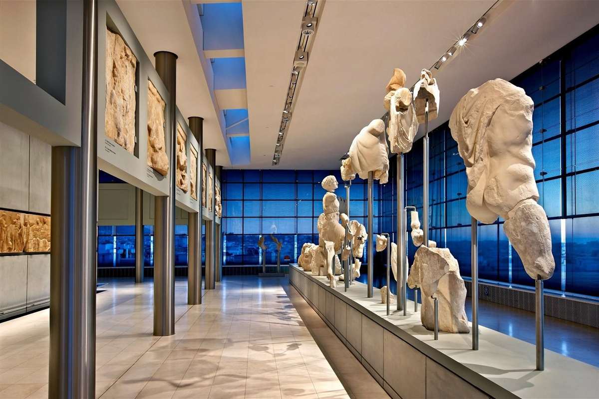 Acropolis guided tour with Acropolis Museum: the Parthenon Gallery with broken marble figures from the pediments standing on a long plinth beside a blue glass wall, and carved metope panels on pillars at the left