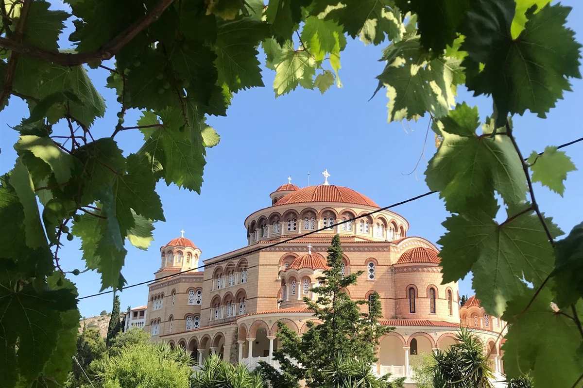 Aegina tours: the large cream-coloured Saint Nektarios monastery with terracotta domes and arched galleries, framed by green vine leaves under a clear blue sky above the island