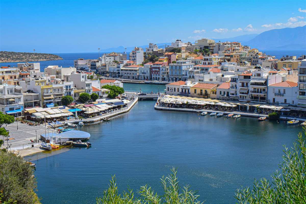 Agios Nikolaos and Spinalonga day trip: the deep blue water of Lake Voulismeni with small boats moored along a quay of white cafe umbrellas, a footbridge, white and terracotta town houses and blue mountains in the distance