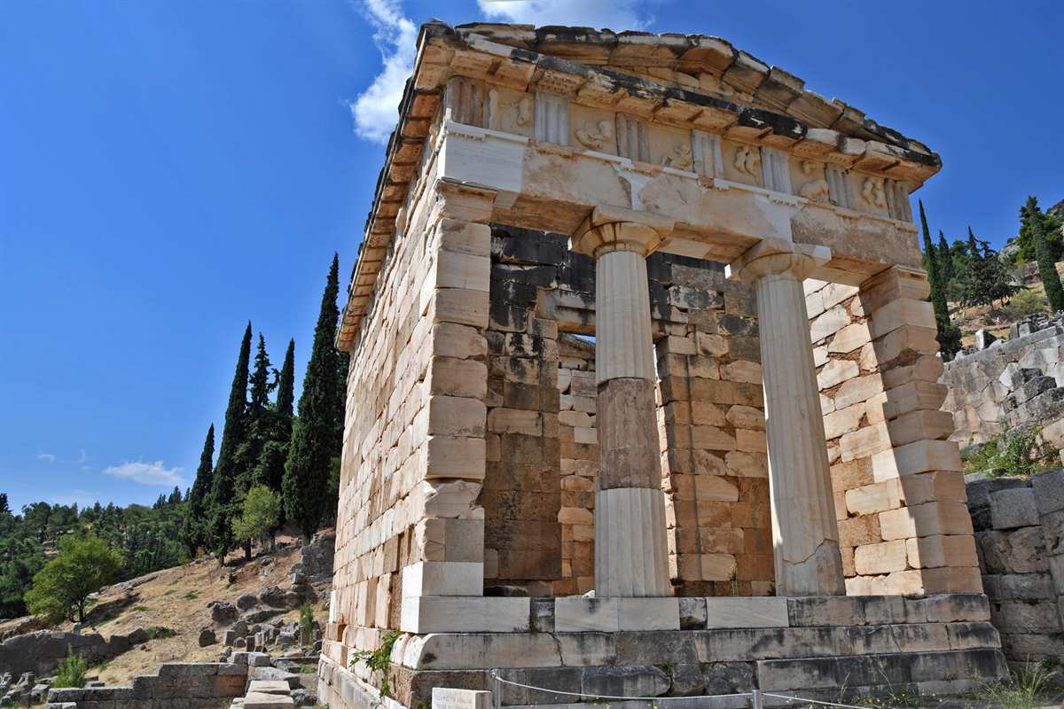 Athens Delphi mythology tour: the Athenian Treasury at Delphi, a small temple of pale stone blocks with two white fluted columns at the front and a carved frieze under its roof, beside tall dark cypress trees under a deep blue sky