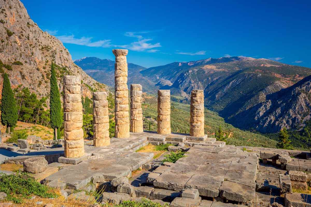 Athens Delphi mythology tour: six tall golden-brown columns of the Temple of Apollo stand on a stone terrace at Delphi, with a pale rock cliff on the left, dark cypress trees, and green mountain valleys under a bright blue sky