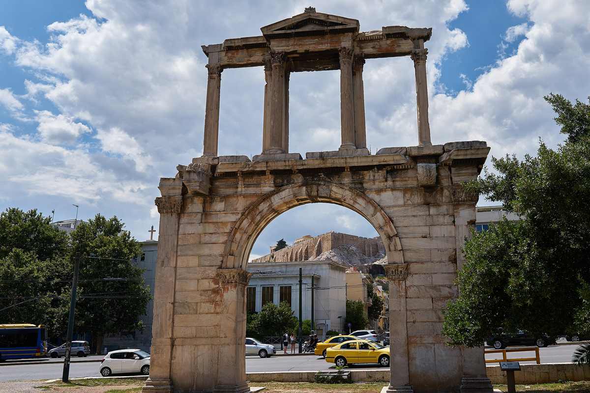 Athens hop on hop off bus tour: the Arch of Hadrian, a pale marble gateway topped by a pediment and four slim columns, with the Acropolis rock and walls seen through its round arch under a blue sky and white clouds