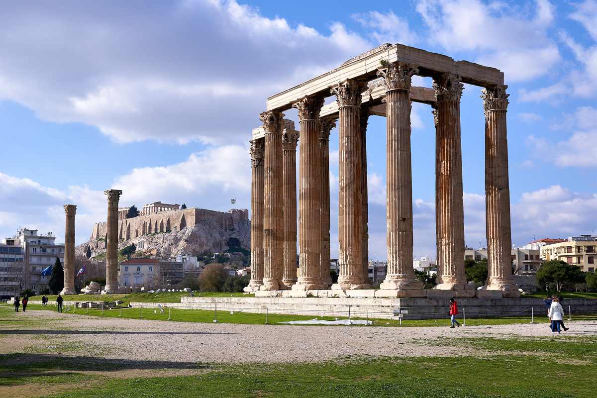 Athens hop on hop off bus tour: the tall fluted Corinthian columns of the Temple of Olympian Zeus in warm honey-coloured stone, with the Acropolis rock and its walls on the hill behind, under a blue sky with white clouds