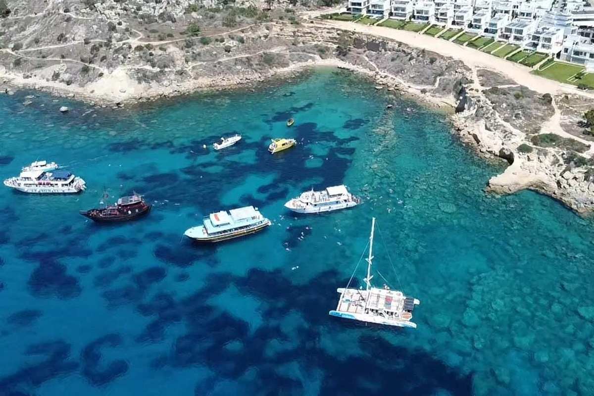 Ayia Napa adults only Blue Lagoon cruise: seen from above, white and wooden tour boats and a white catamaran at anchor in a deep blue and turquoise bay, with dark reef patches in the clear water and a pale rocky shore with white houses along the top