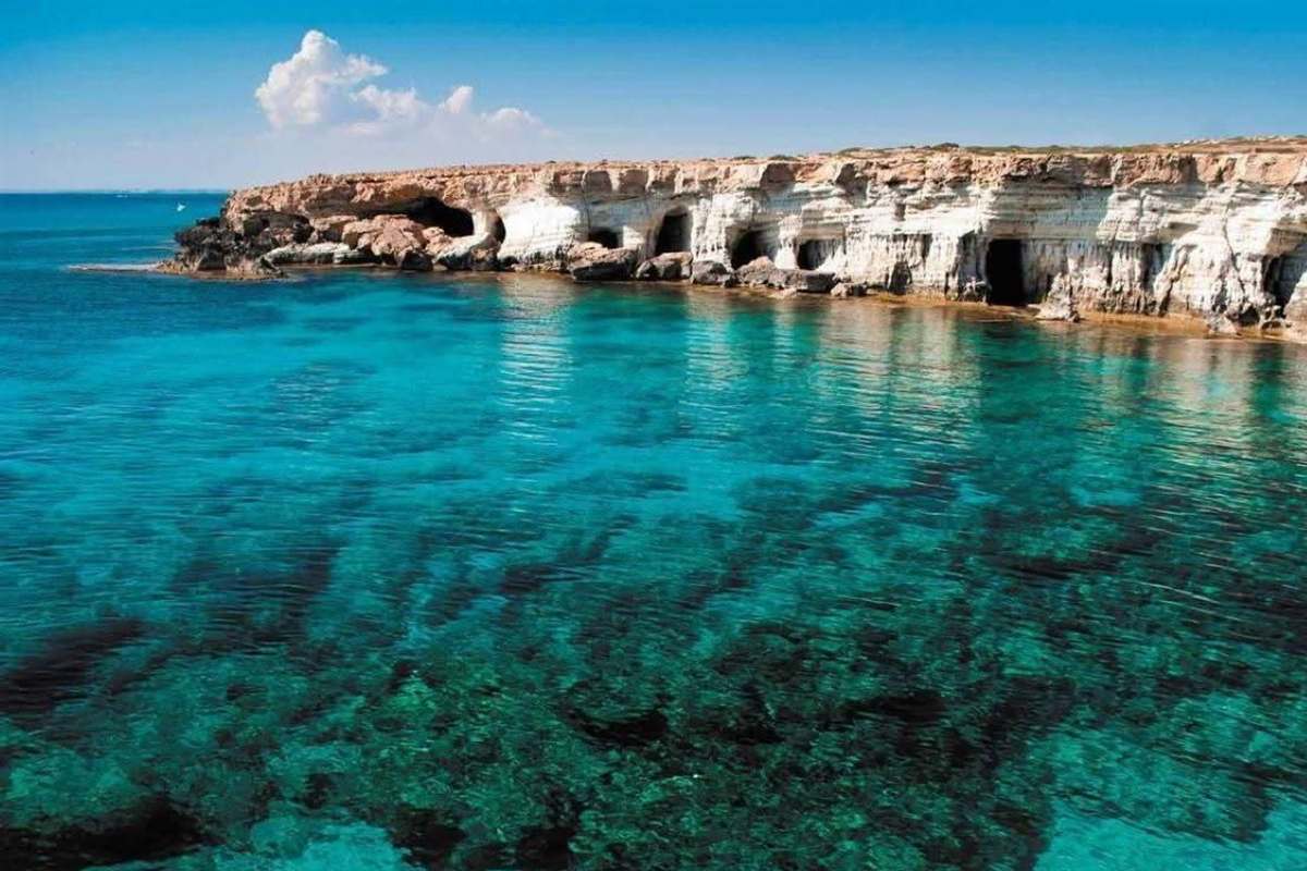 Ayia Napa adults only Blue Lagoon cruise: pale limestone cliffs with dark sea cave openings along the Cape Greco shore, seen from the water over clear turquoise and green shallows with dark reef patches under a bright blue sky and one white cloud
