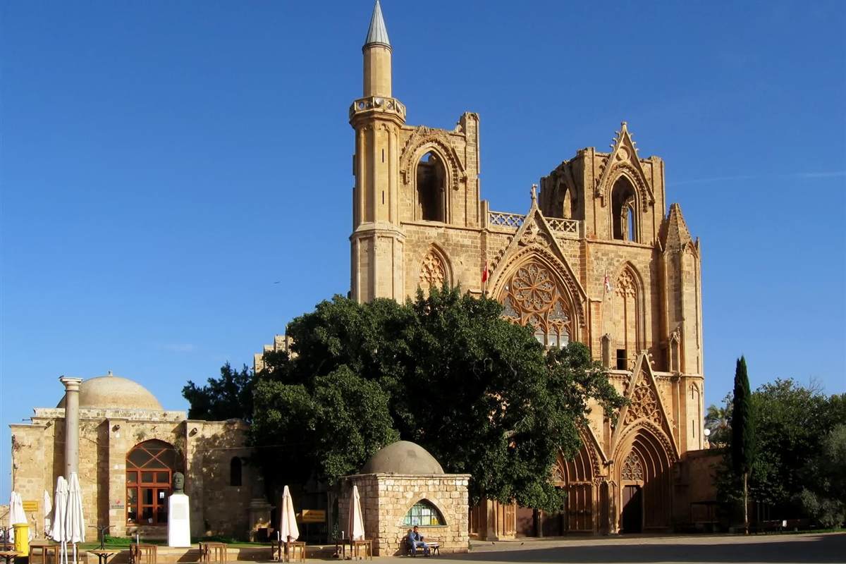 Ayia Napa Famagusta ghost town tour: the Gothic west front of the former Saint Nicholas Cathedral in Famagusta, now the Lala Mustafa Pasha Mosque, in honey-coloured stone with a tall minaret, a large rose window and a big green tree under a clear blue sky