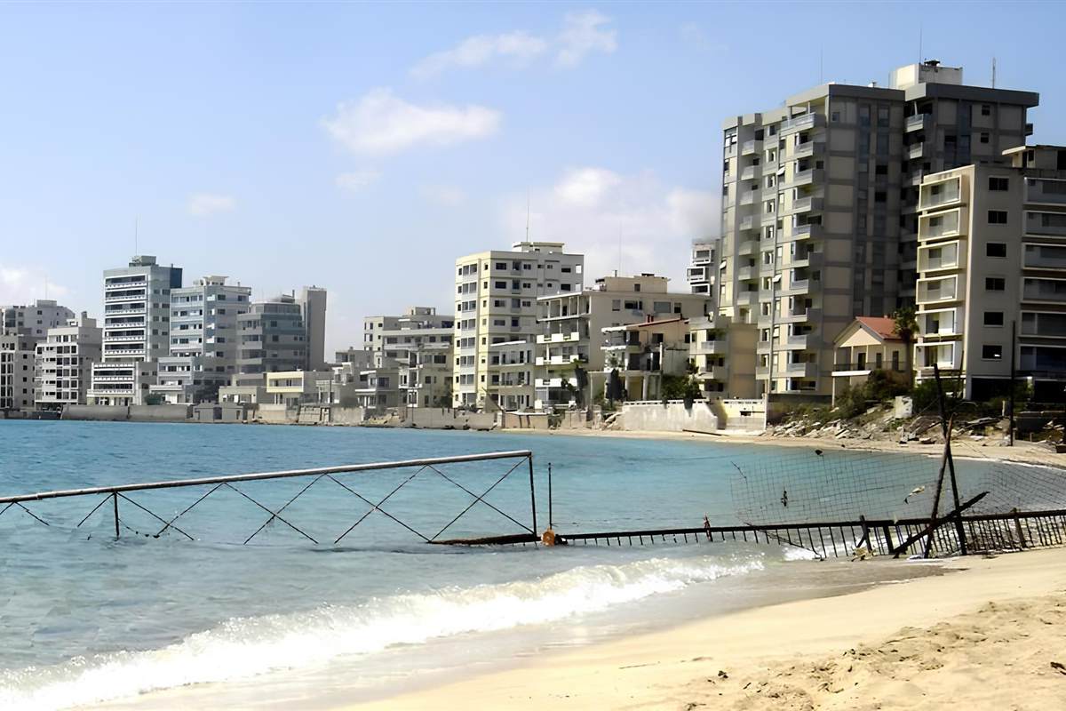 Ayia Napa Famagusta ghost town tour: a row of tall pale apartment and hotel blocks of Varosha above a sandy beach, with a rusty fence running into the calm blue sea under a light blue sky