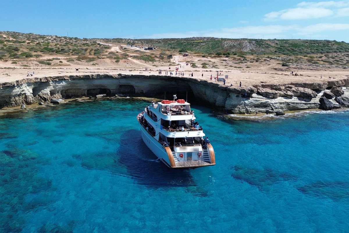 Ayia Napa lazy day cruise: a white multi-deck cruise boat with orange safety gear on its top deck floating in clear turquoise water below pale limestone sea caves on the Cape Greco coast, with visitors on the clifftop under a bright blue sky