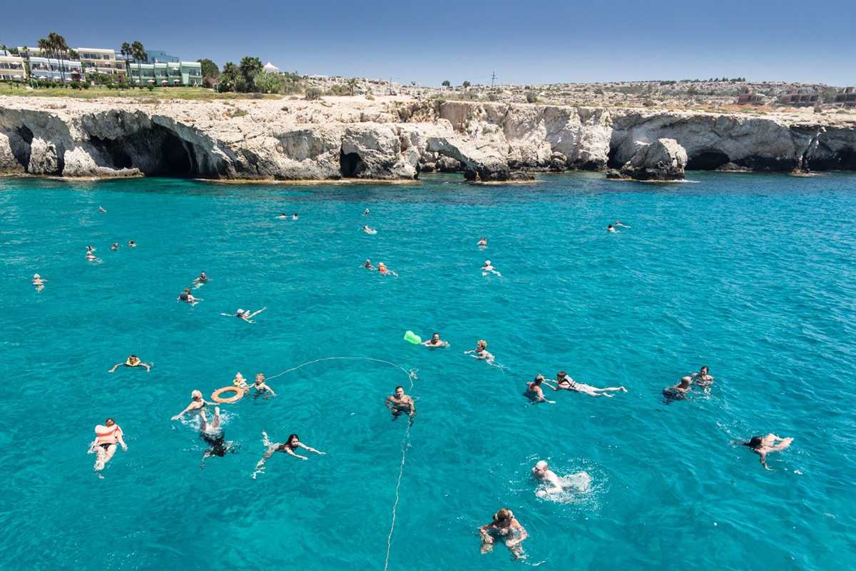 Ayia Napa lazy day cruise: swimmers spread across clear turquoise water below pale limestone sea cliffs with dark cave openings, a hotel garden with palm trees on the clifftop and a deep blue sky