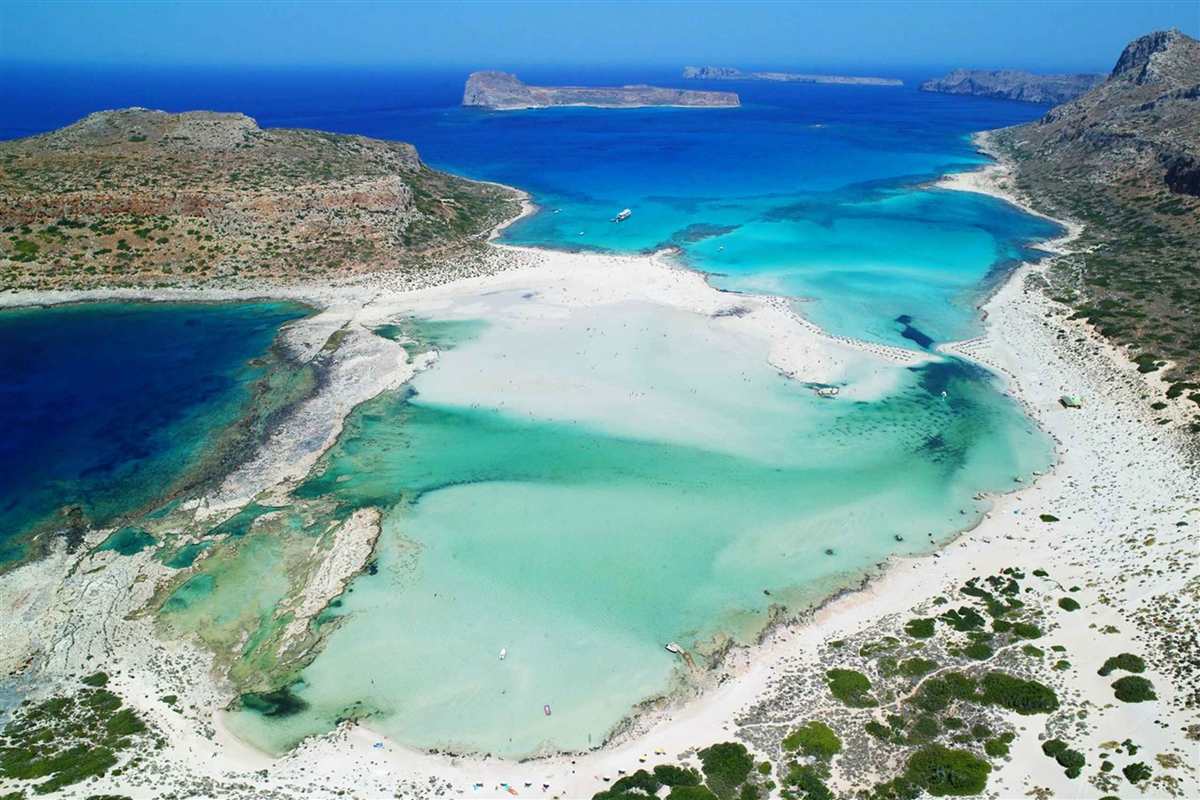 Balos and Gramvousa boat tour from Chania: an aerial view of the Balos lagoon with pale sand bars and shallow turquoise water, deep blue sea beyond, and the rocky island of Gramvousa on the horizon under a clear sky