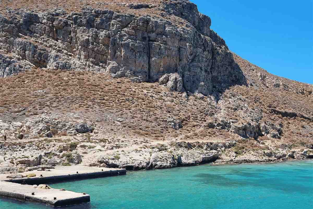 Balos and Gramvousa boat tour from Chania: a steep grey and brown rocky slope on the Gramvousa coast above a concrete stone jetty and calm turquoise water under a clear blue sky