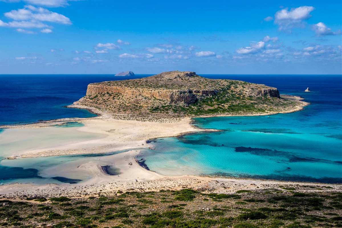 Balos and Gramvousa catamaran trip: the flat-topped Tigani headland above the Balos lagoon, with pale sand bars, turquoise shallows, deep blue open sea and a small rocky islet on the horizon under a blue sky with white clouds