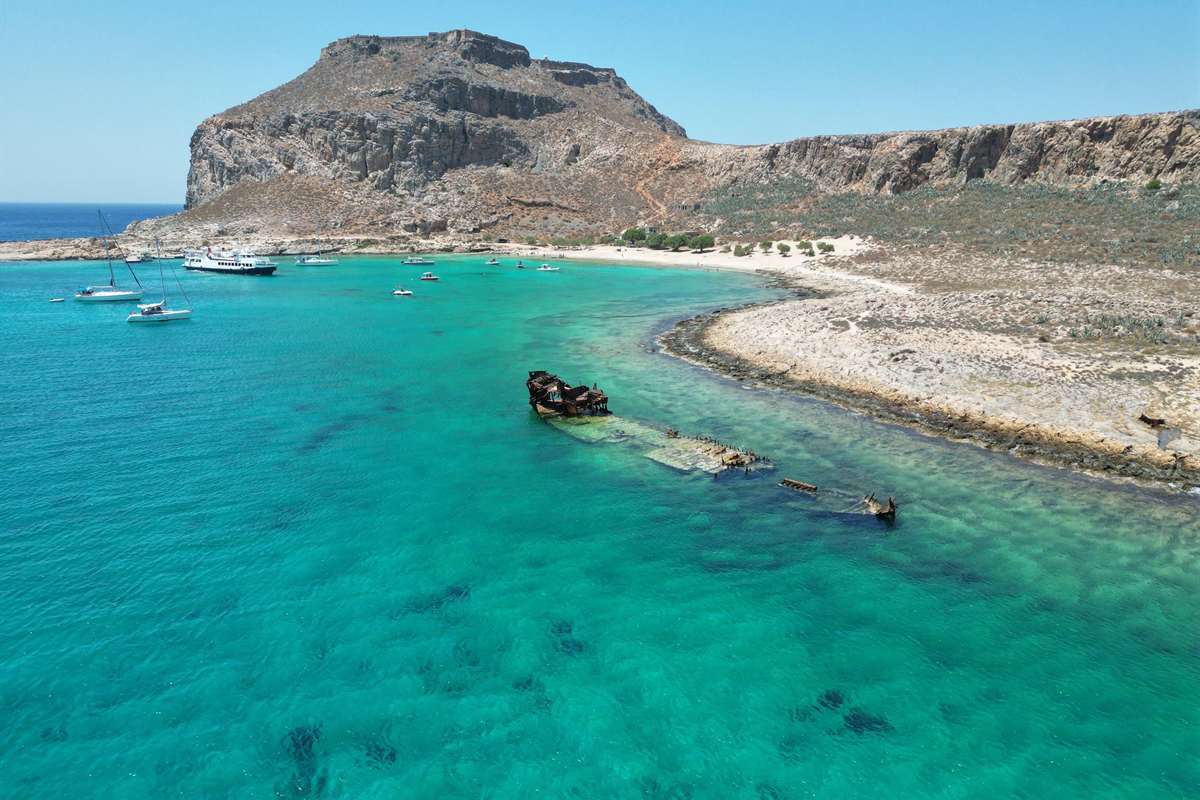 Balos and Gramvousa catamaran trip: the rocky hill of Imeri Gramvousa with the walls of the Venetian fortress along its summit, above a turquoise bay with white sailing boats and a passenger boat at anchor and a rusty shipwreck lying in the shallows
