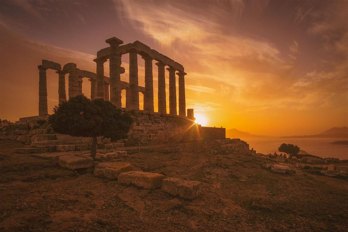 Cape Sounion sunset tour from Athens: the long colonnade of the Temple of Poseidon in warm orange light on the cliff edge, a dark green tree below the columns and a low sun above the sea under swirling orange and purple clouds