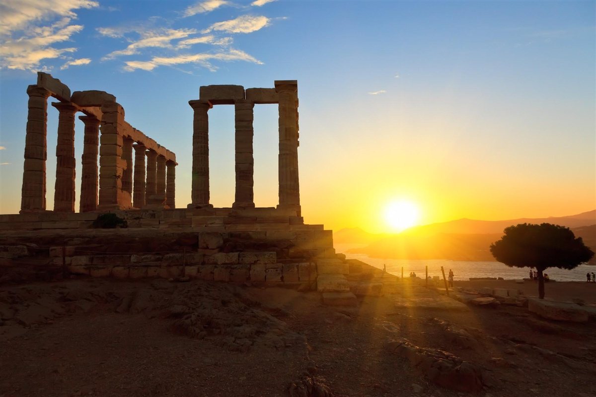 Cape Sounion sunset tour from Athens: the weathered Doric columns of the Temple of Poseidon stand dark against a blue sky above a glowing yellow setting sun, with a lone pine tree on the right and a hazy mountain coast beyond the calm sea