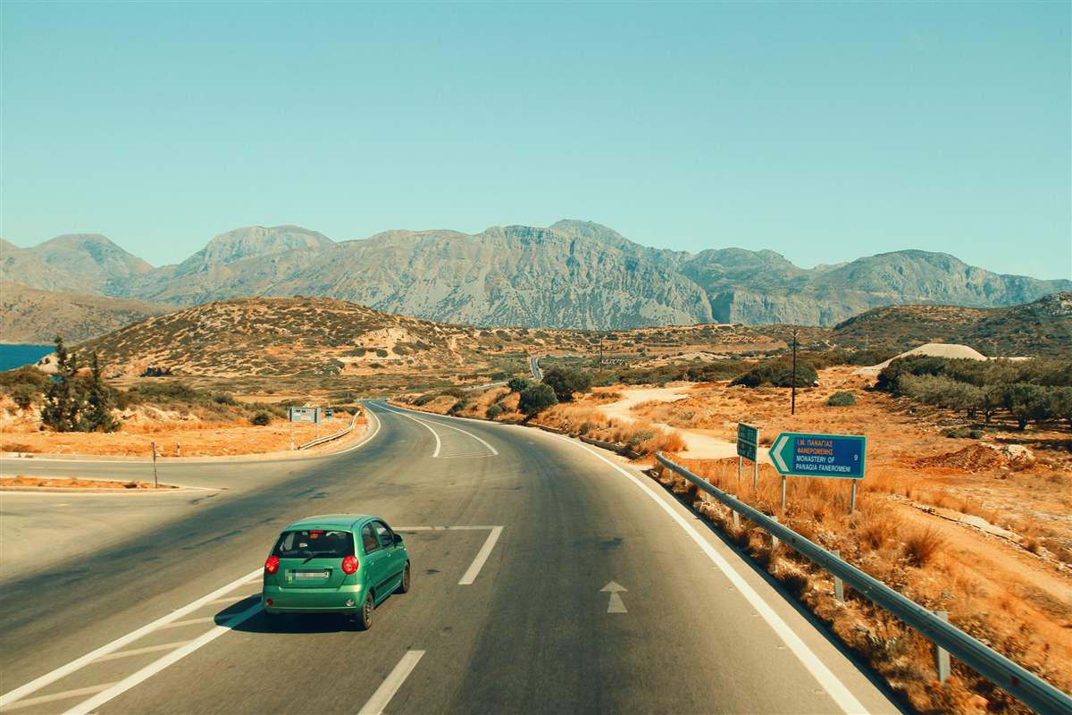 Car rental Greece: a small green hatchback drives along a curving mountain road in Crete past a blue road sign, with bare brown hills and a clear sky ahead.