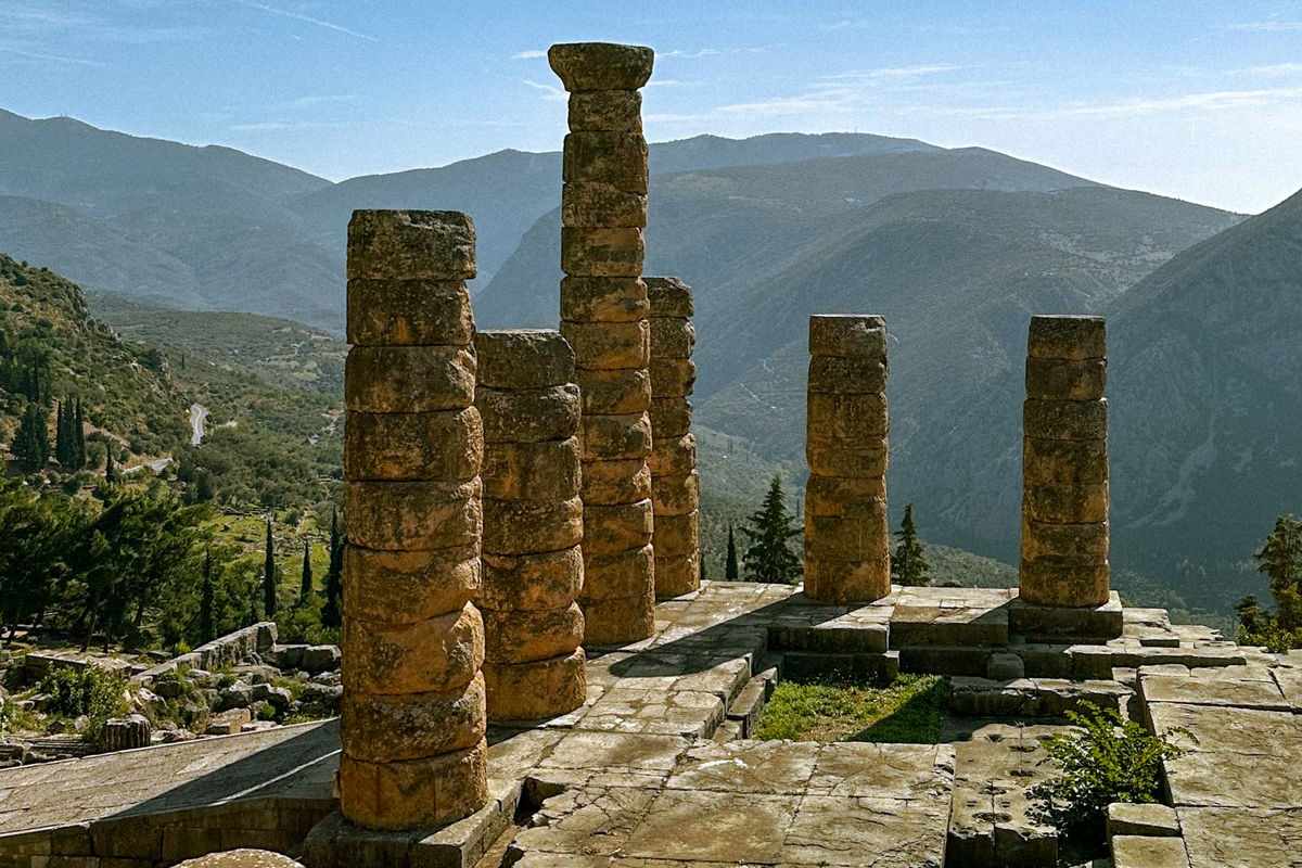 Classical tours Greece: the stacked stone drums of the Temple of Apollo at Delphi rise against a pale blue sky above the mountains of the valley below.