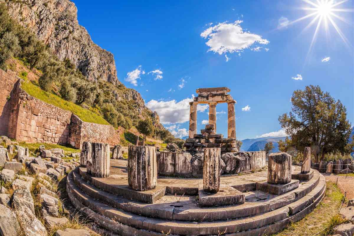 Delphi tours: the round Tholos at Delphi, three standing columns above a ring of stone drums and a low circular base, under a bright blue sky with the sun in the corner