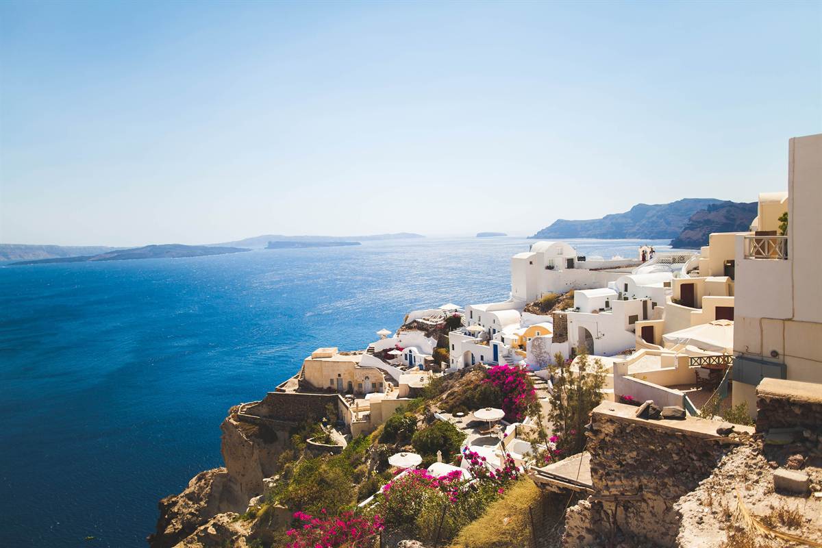Greece tours: whitewashed and cream-coloured houses with pink bougainvillea on the caldera cliff at Oia on Santorini, above deep blue water with dark volcanic islands on the horizon under a clear sky