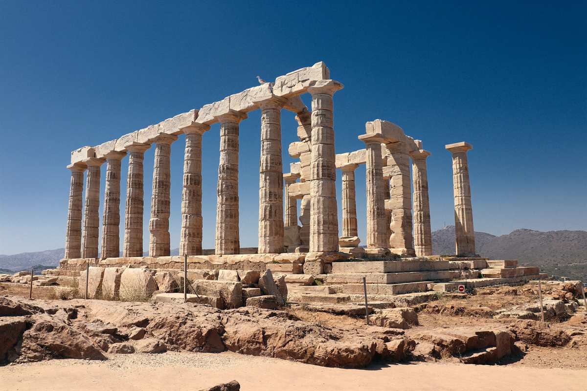 Greece tours: the ruined Temple of Poseidon at Cape Sounion, two rows of tall weathered Doric marble columns carrying stone beams, standing on rocky ground under a deep blue cloudless sky