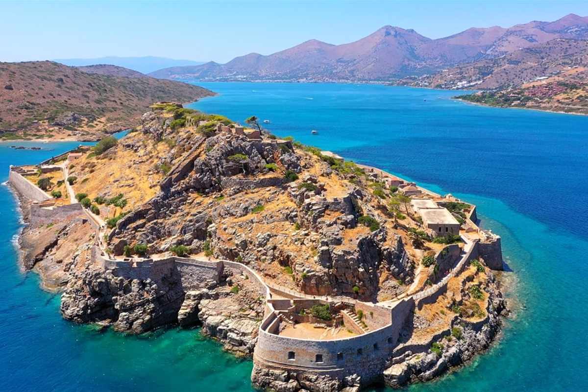 Greek islands cruises: the walled fortress island of Spinalonga, with stone bastions and dry scrub on its hill, set in deep blue and turquoise water with mountains on the far shore