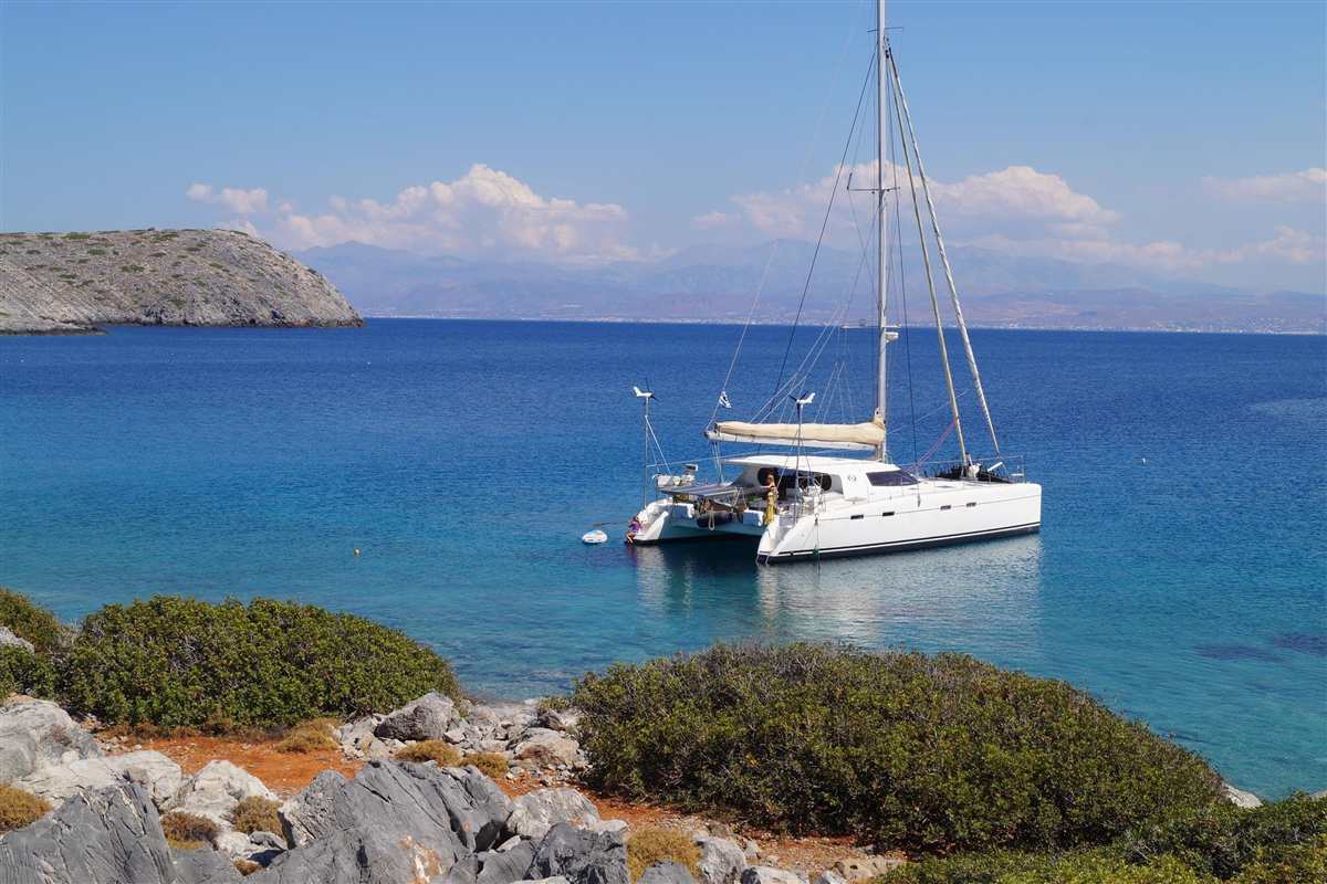 Heraklion Dia Island sailing cruise: a white sailing catamaran at anchor in calm deep-blue water beside a rocky shore with green shrubs, a low rocky headland on the left and the hazy mountains of Crete under a blue sky with white clouds