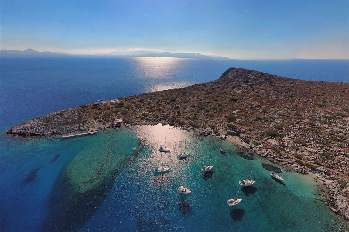 Heraklion Dia Island sailing cruise: an aerial view of white sailing boats anchored in a turquoise bay on the rocky, scrub-covered island of Dia, with a small jetty and two white buildings on the shore and the Cretan coast on the horizon