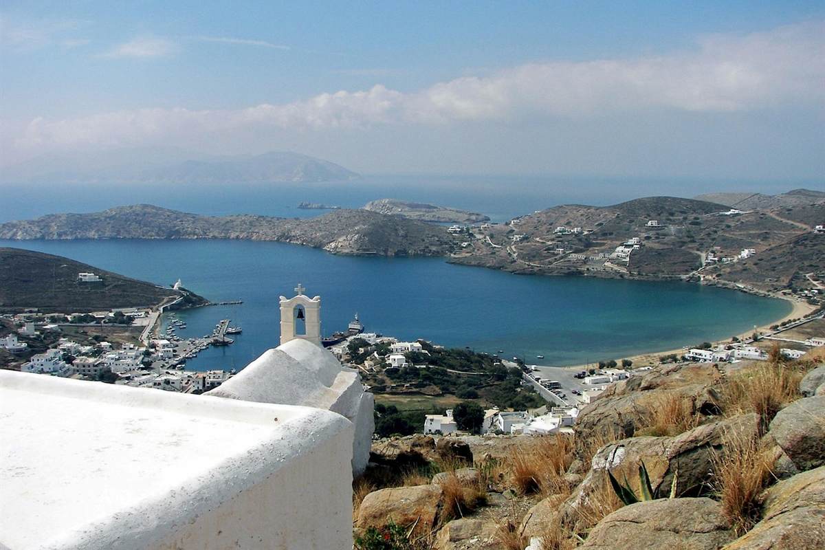 Ios tours: a bay of deep turquoise water with a white harbour village and a whitewashed bell tower in the foreground, boats moored at the quay and rocky hills behind