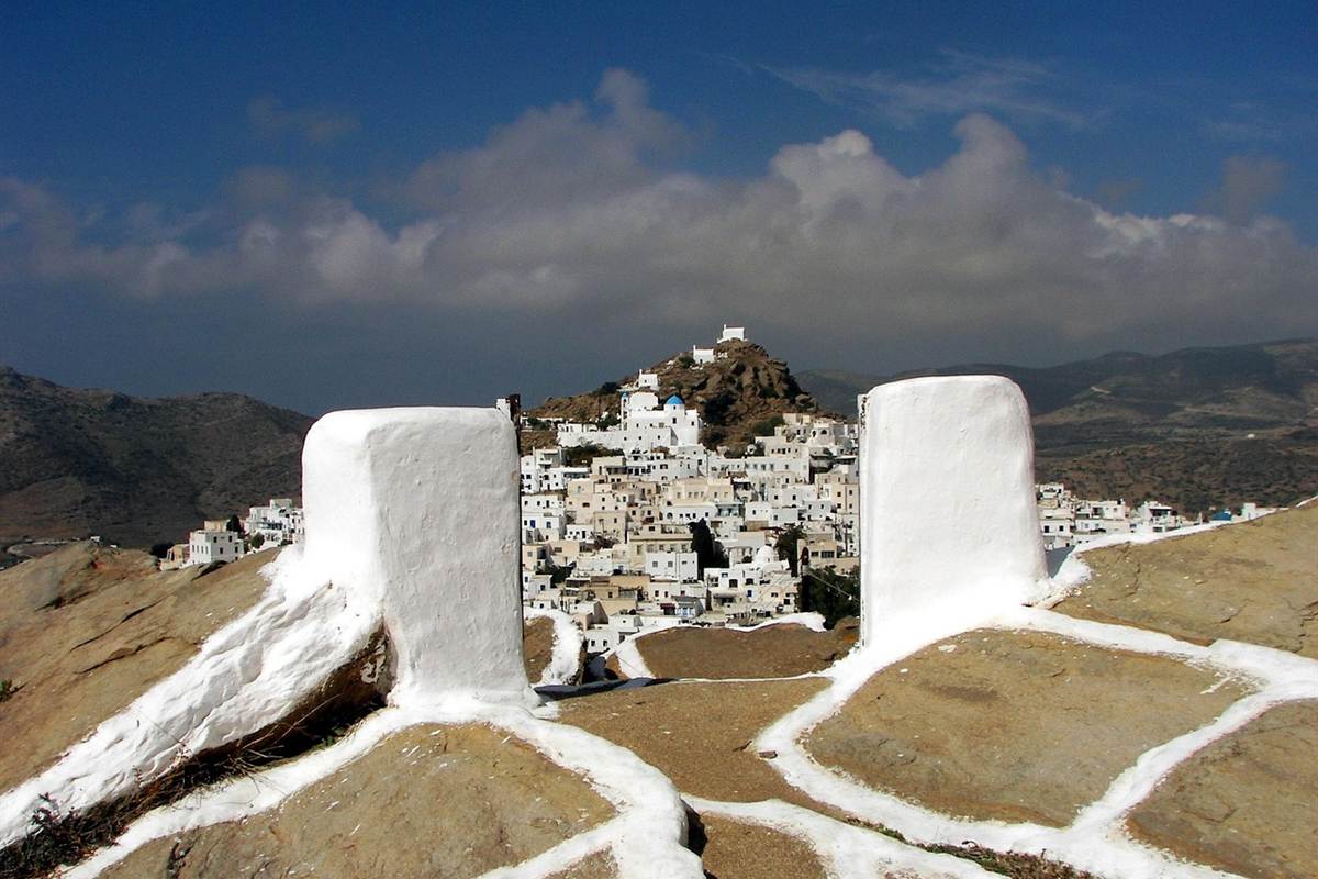 Ios tours: the white houses of Chora stacked on a rocky hill, with a blue-domed chapel and a small white chapel on the summit under a deep blue sky, dry hillside and mountains beyond