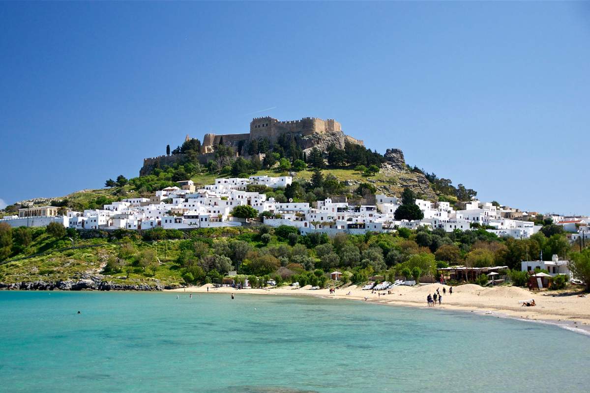 Lindos and Seven Springs bus tour from Rhodes: the walled Acropolis of Lindos on its rocky green hill above the white village houses and a curved sandy bay with turquoise water, under a clear blue sky