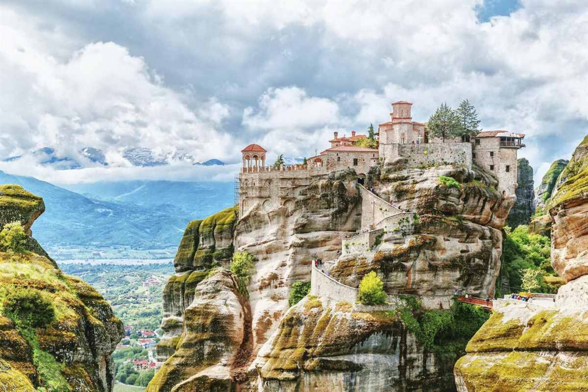 Meteora day trip from Thessaloniki: a stone monastery with red-tiled roofs, a small red-roofed lookout and a square bell tower on top of a mossy golden rock cliff, with a green valley, blue mountains and white clouds behind