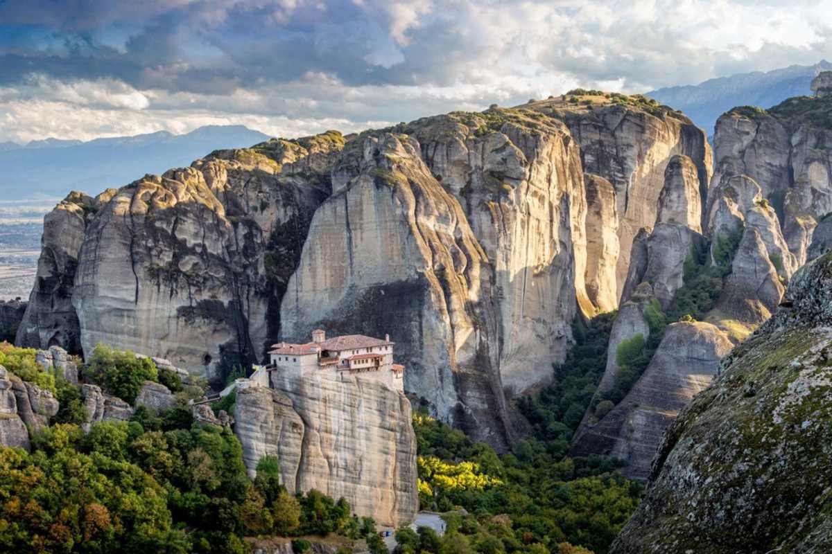 Meteora day trip from Thessaloniki: the pale stone Roussanou monastery with red-tiled roofs on a narrow rock pillar below huge golden sandstone cliffs, with green trees, distant mountains and a cloudy sky behind