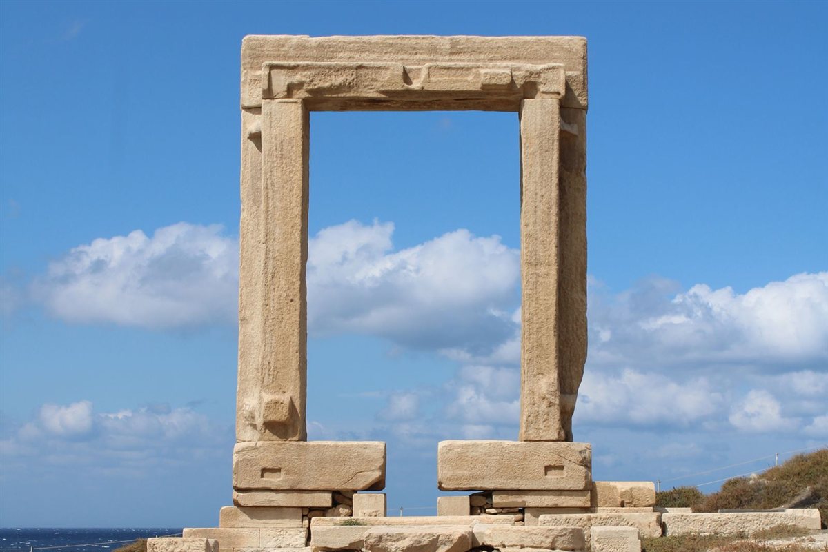 Naxos tours: the Portara, the marble doorway of the unfinished Temple of Apollo on Naxos, stands whole under a clear blue sky with dry grass and fallen marble blocks around its base