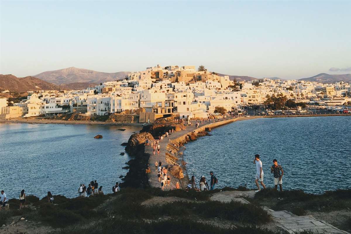 Naxos tours: Naxos Town and its white houses on the hill beneath the Kastro, seen from the causeway to Palatia at golden hour, with people on the stone path and clear sky over the sea