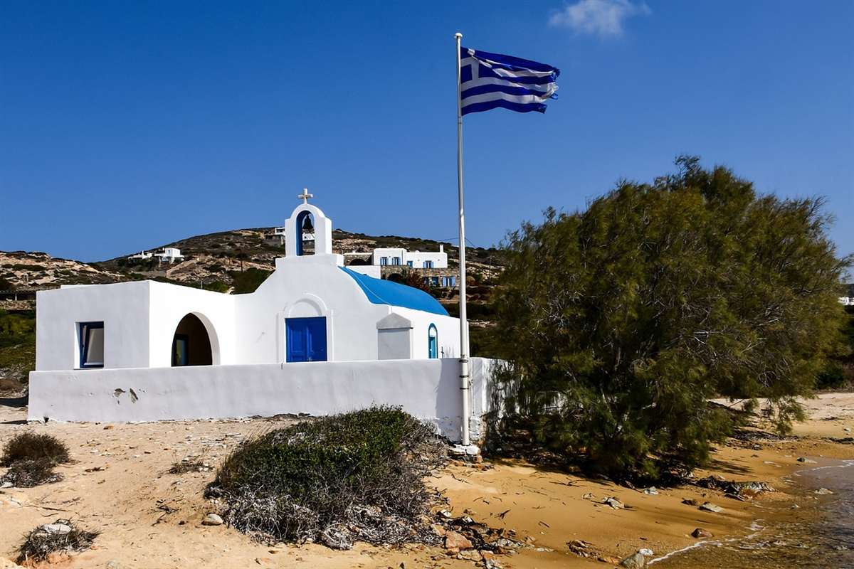 Paros tours: a white chapel with a blue dome, blue doors and a Greek flag on a sandy Antiparos beach under a clear blue sky, with a tamarisk tree beside it