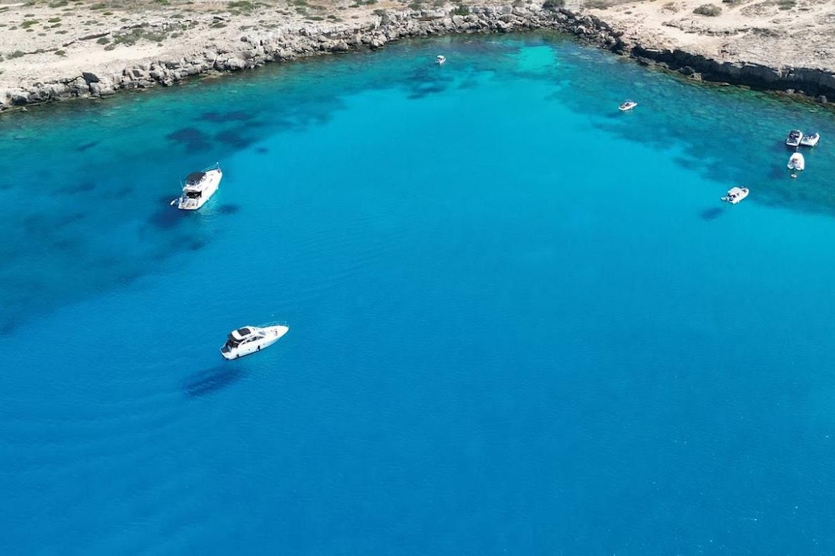 Protaras Blue Lagoon cruise: white motor boats at anchor in a deep blue and turquoise cove with a pale rocky limestone shore along the top of the bay and dark reef patches in the clear water