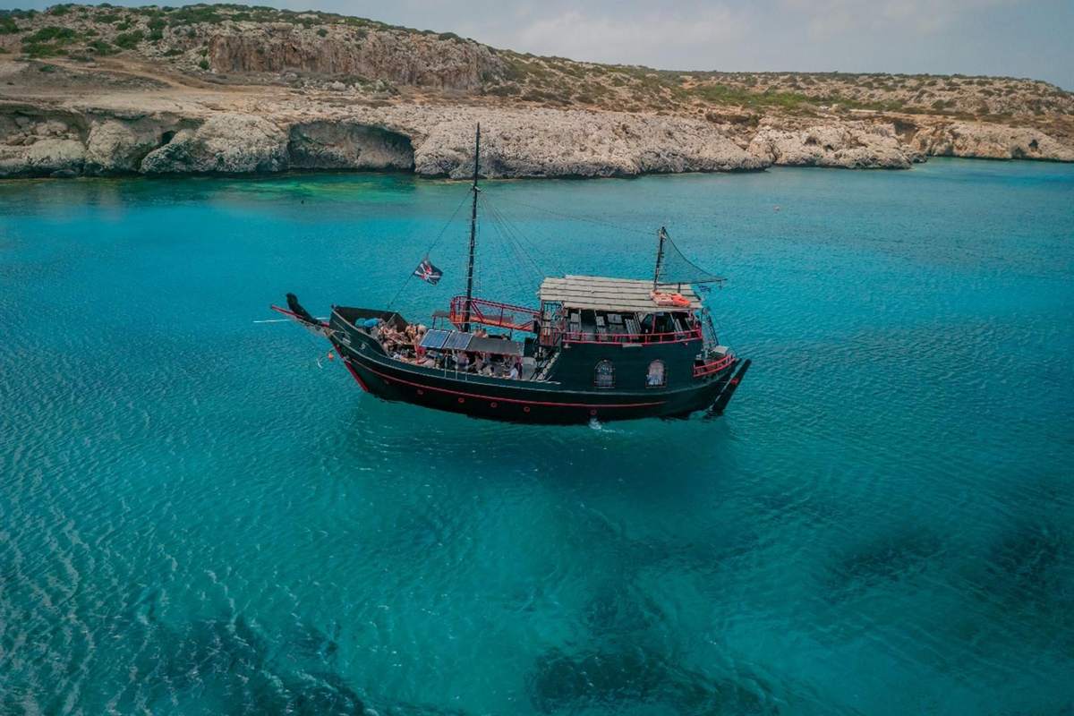 Protaras Blue Lagoon cruise: a black wooden two-masted tour boat with a small flag and a red-trimmed deck floats on bright turquoise water below low pale limestone cliffs on the Cape Greco coast