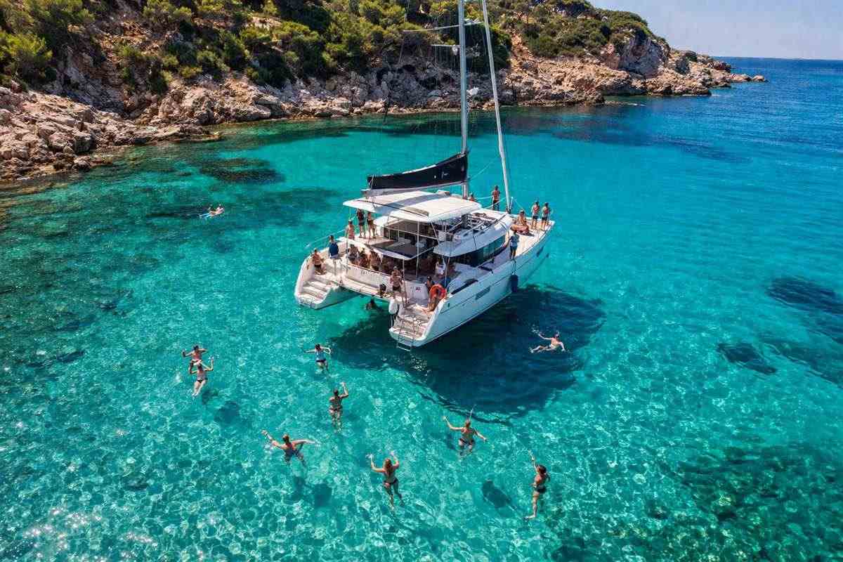 Rhodes all inclusive catamaran cruise: a white catamaran with a dark furled mainsail anchored in clear turquoise water beside a rocky pine-covered shore, with swimmers floating around the hulls