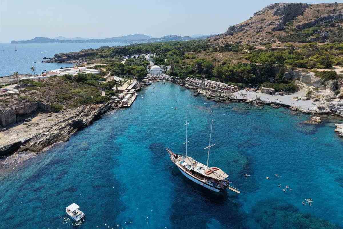 Rhodes all inclusive catamaran cruise: aerial view of Kallithea Springs with the white domed rotunda at the head of a long blue inlet, sunbeds along the shore, a two-masted wooden sailing boat at anchor and swimmers in the water below a rocky green hill