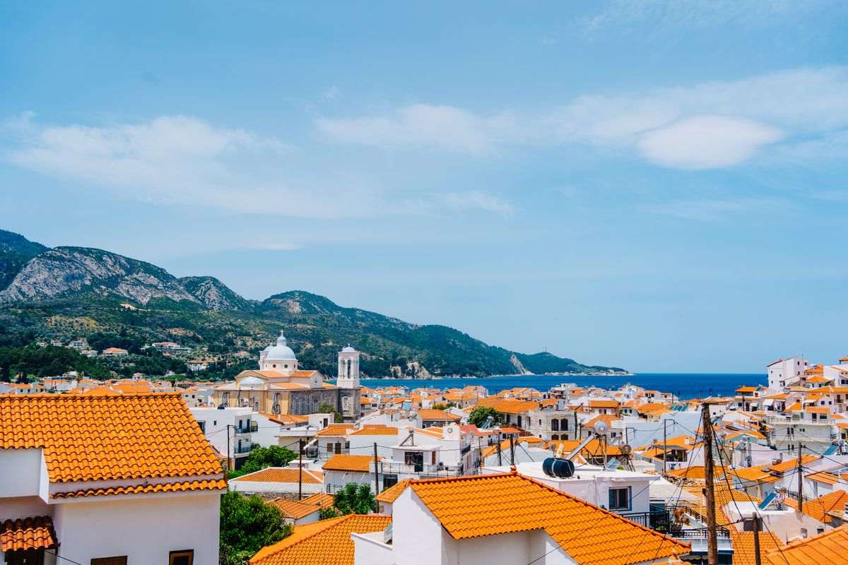 Samos tours: the orange-roofed village of Kokkari spread above the blue bay, with a domed church, green mountains and a clear blue sky