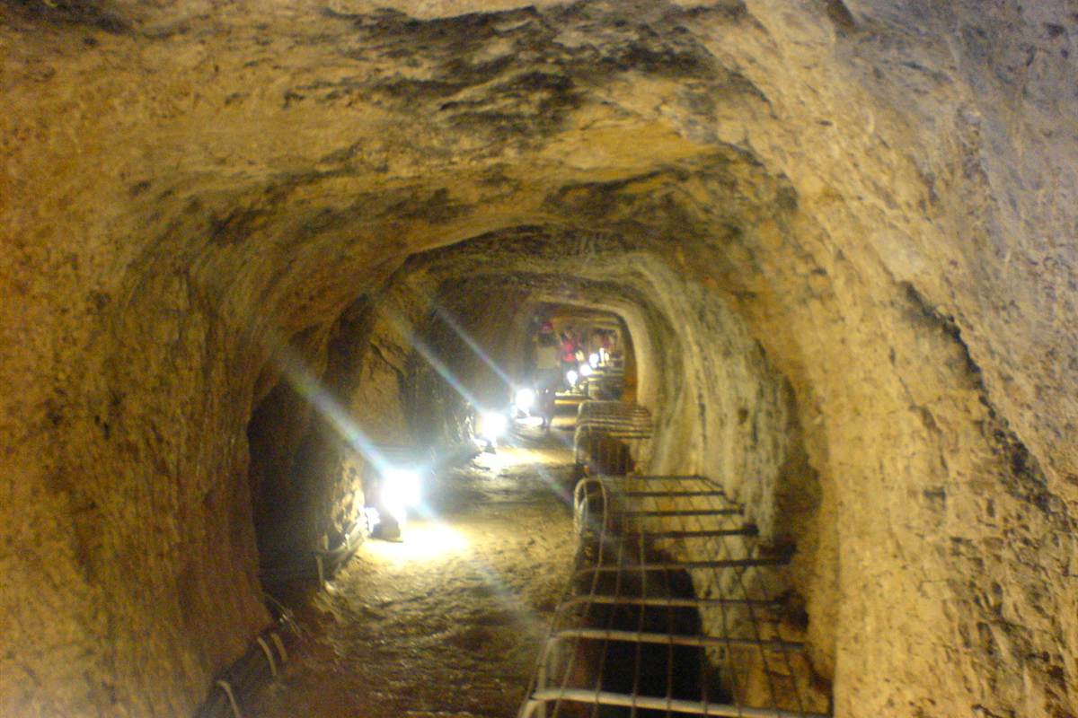 Samos tours: a lit stone tunnel of the Tunnel of Eupalinos with rounded rock walls, a metal walkway along the right side and small lights far ahead