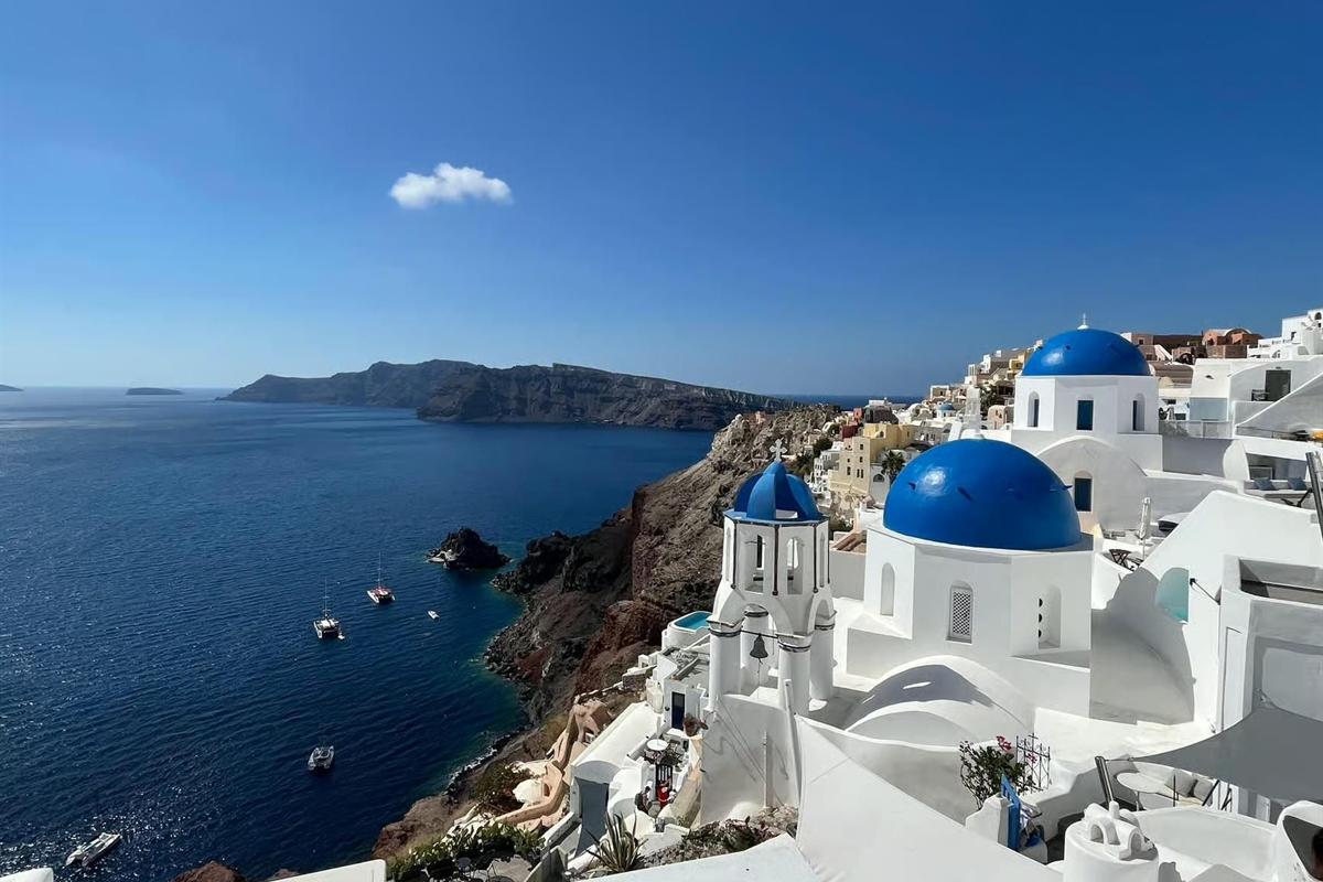 Santorini flying dress photoshoot: white houses and blue domes with a white bell tower on the caldera rim at Oia, deep blue water below with sailing boats and a dark cliff headland across the bay under a clear blue sky