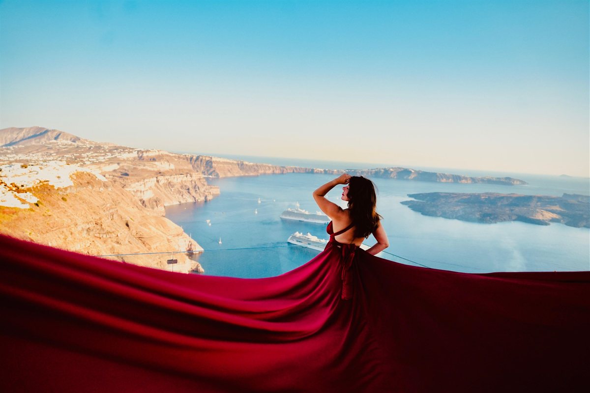 Santorini flying dress photoshoot: a woman seen from behind with one hand raised to her hair, a long dark red dress spread in a wide train across the foreground above the caldera, with golden cliffs, white villages, two cruise ships and a dark volcanic island behind her under a pale blue sky
