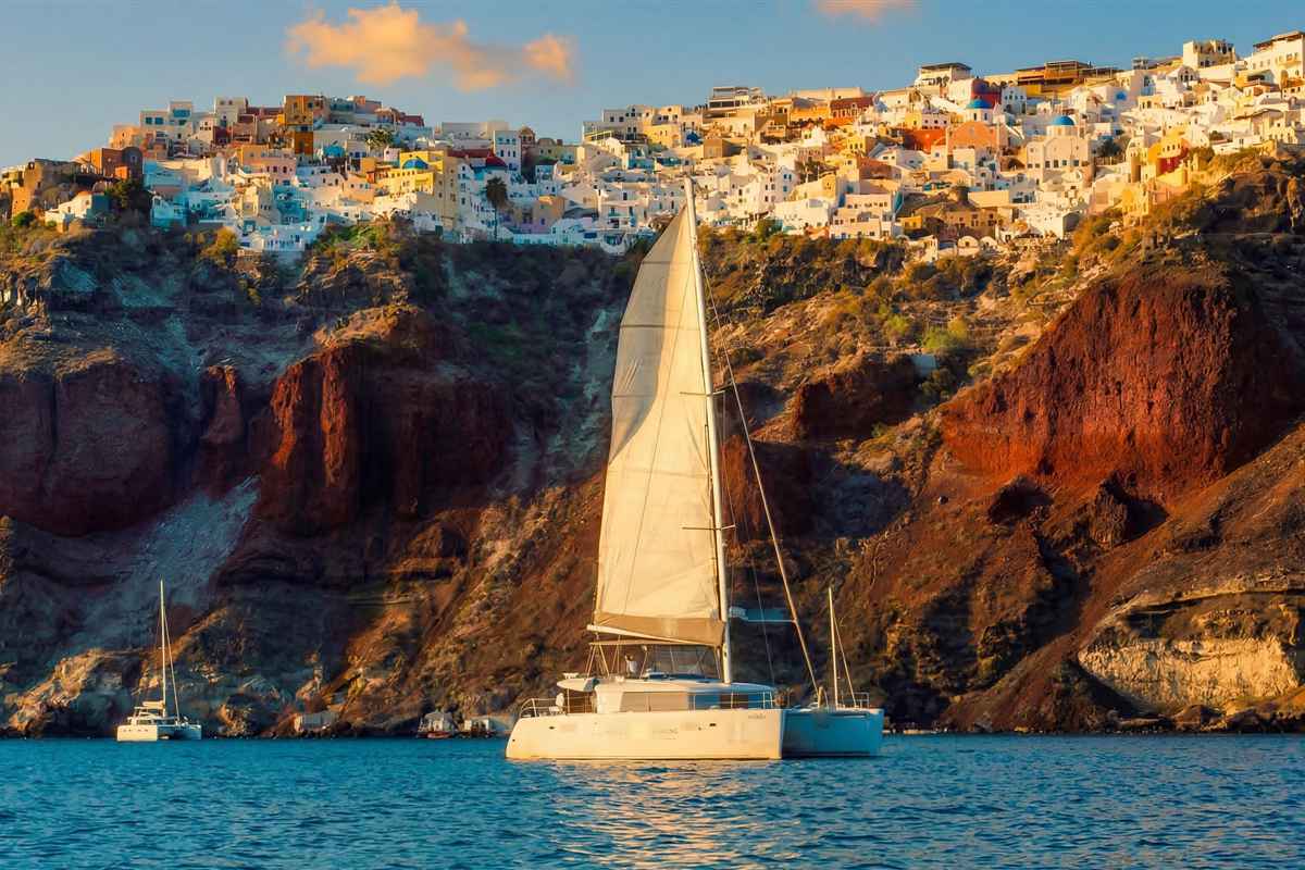 Santorini Thirasia and Oia sunset cruise: a white sailing catamaran with a cream mainsail on calm blue water below the red and brown caldera cliffs, with the white and pastel houses of Oia along the rim in warm evening light