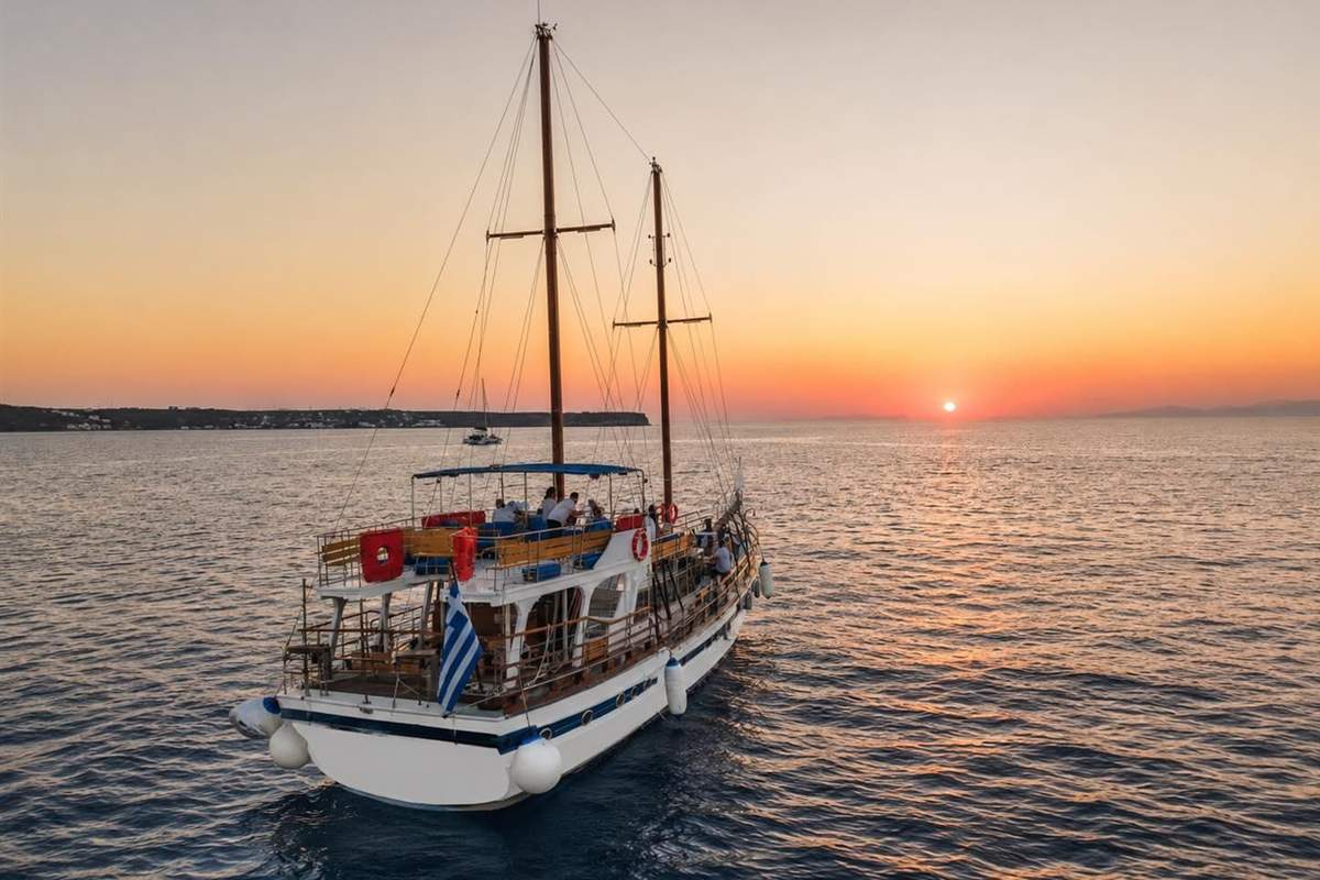 Santorini Thirasia and Oia sunset cruise: a two-masted white and wooden cruise boat with a Greek flag at its stern on calm water while the orange sun sets over the horizon, with a low island coast on the left