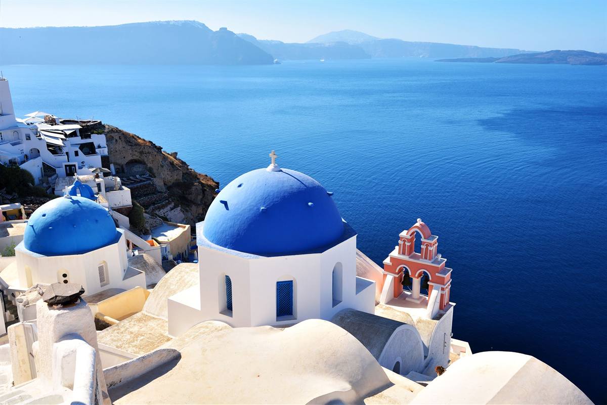 Santorini top sights day trip: a white church with a bright blue dome and a small pink and red bell tower on the cliff edge at Oia, a second blue dome beside it, above deep blue caldera water with the far rim of the island under a pale sky