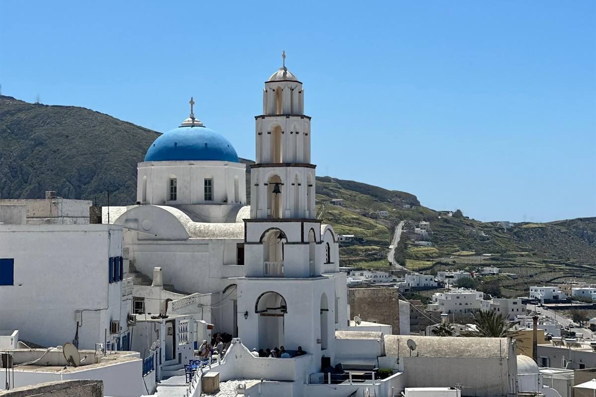 Santorini top sights day trip: a white church with a blue dome and a tall white bell tower with three tiers of arched openings in the village of Pyrgos, green hills behind and a clear blue sky