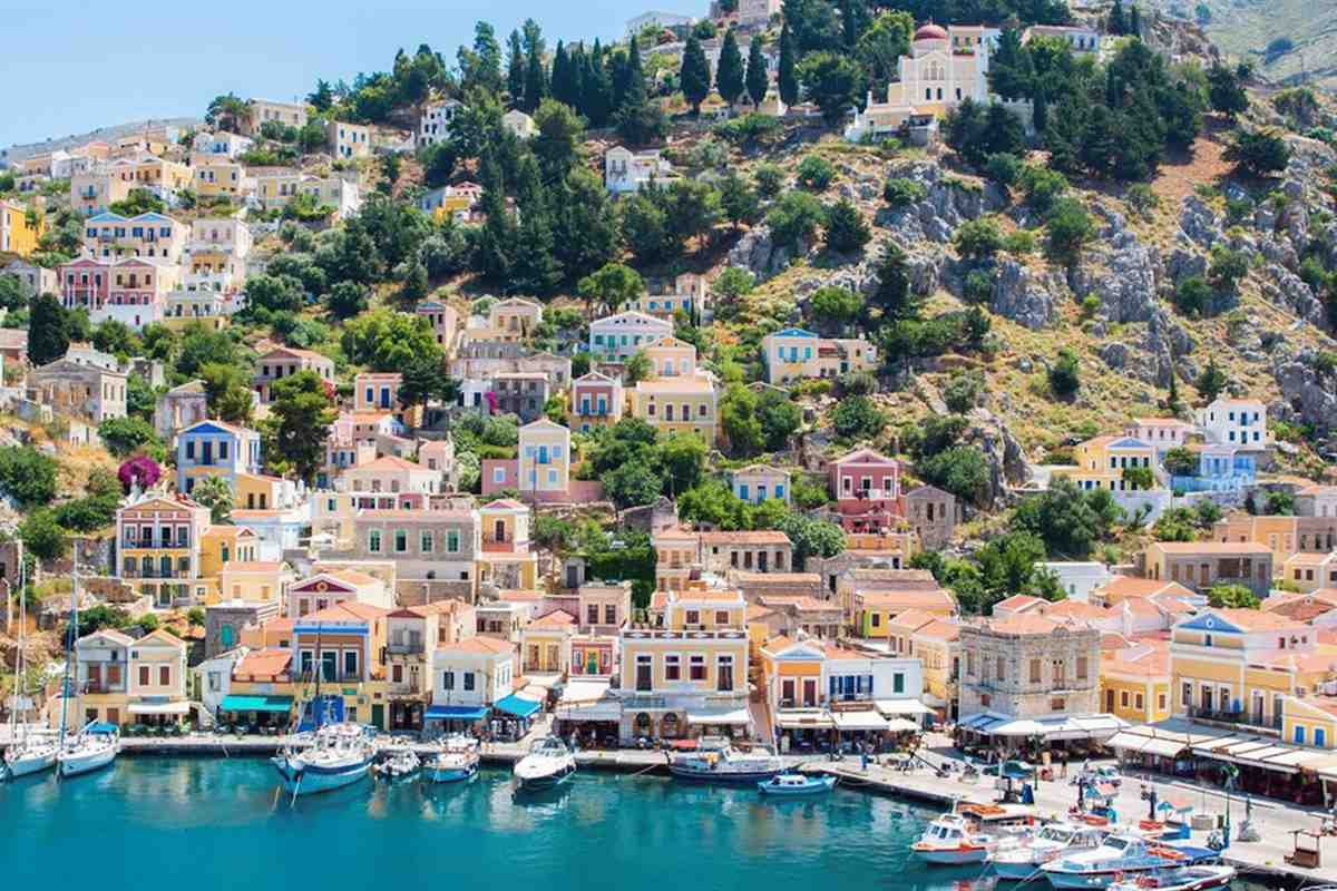 Symi day trip from Rhodes: pastel yellow, pink and white neoclassical houses stacked up the green and rocky hillside above Gialos harbour, with moored sailing boats and fishing boats on the turquoise water below and a red-domed church on the ridge
