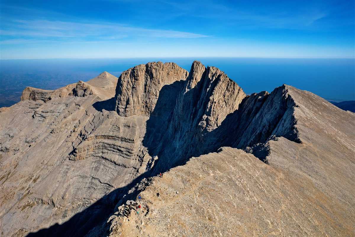 Thessaloniki Dion and Mount Olympus day trip: the pale grey limestone summit ridge of Mount Olympus with the rock tower of Mytikas, sunlit slopes and deep shadow in the gullies, and a flat blue haze of sea and plain far below under a clear blue sky