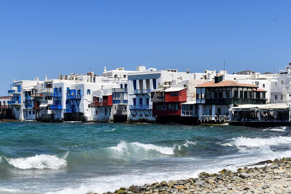 weddings in greece: a seaside row of whitewashed houses with blue balconies and a red-shuttered house on the edge of the Aegean in Little Venice, Mykonos, under a clear blue sky